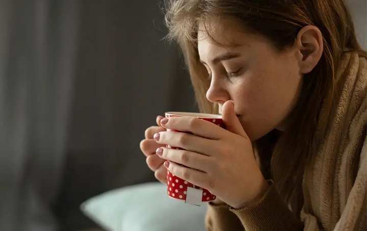 A woman taking a sip of a hot beverage while sick during the holidays.