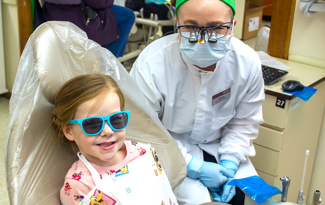 Smiling girl with shades in dental chair with hygienist/dentist sitting next to her in work protective gear